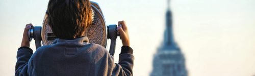 A child uses binoculars to view the Empire State Building in New York City at sunset.